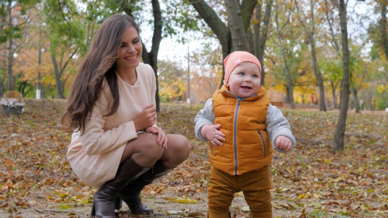 Mama podstiče bebu koja pravi prve korake u jesenjem parku