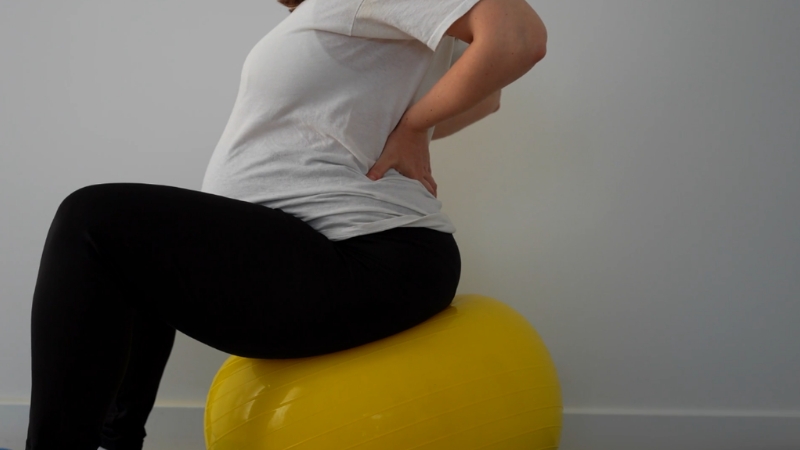 Trudnica sedi na pilates lopti i drži se za leđa zbog bola u leđima u trudnoći